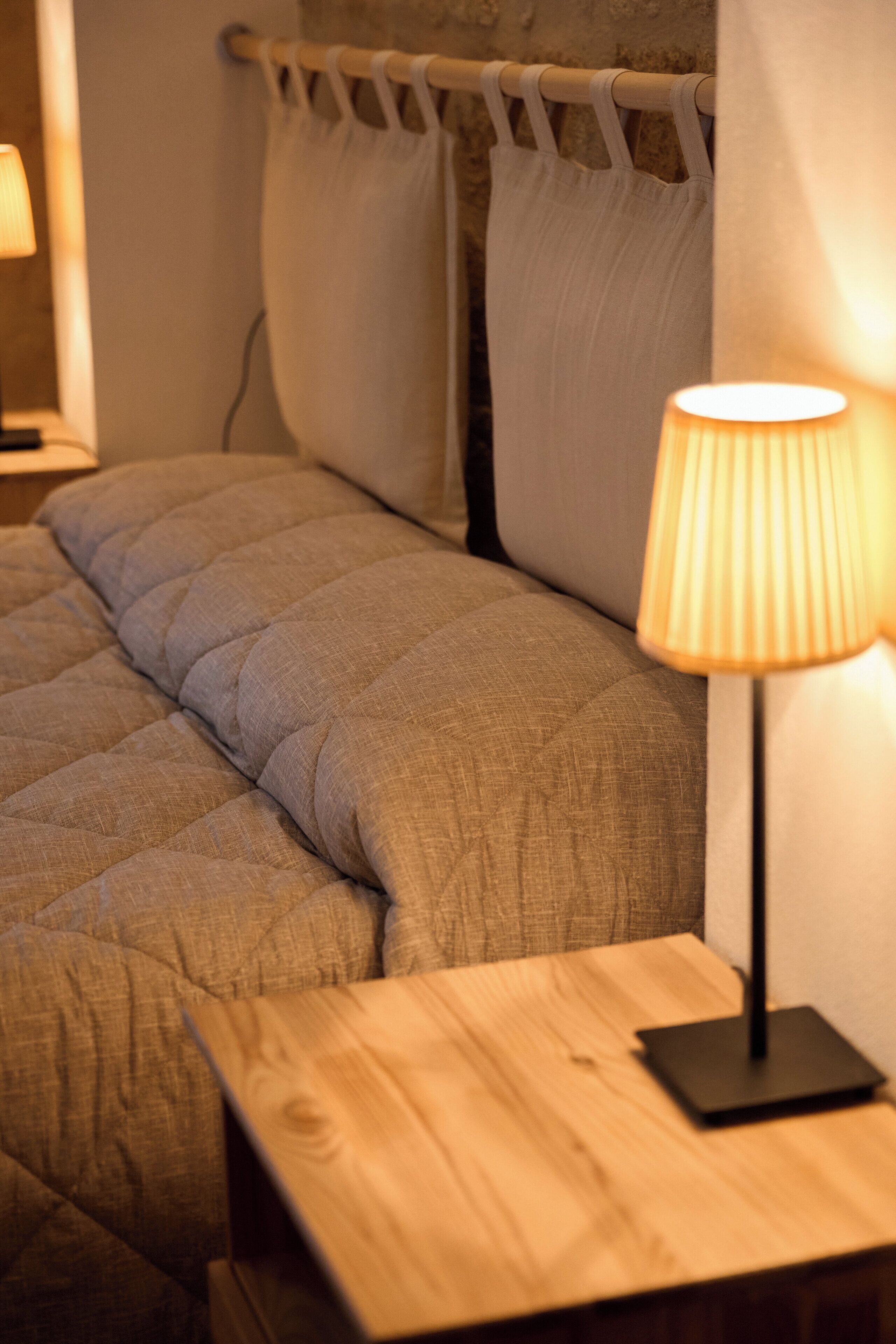 Una cama bien hecha con un edredón acolchado, una lámpara de noche y una mesita de madera.