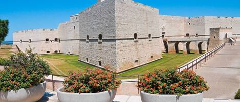 Una fortaleza de piedra histórica con una torre central, rodeada por un camino pavimentado y grandes macetas con flores.