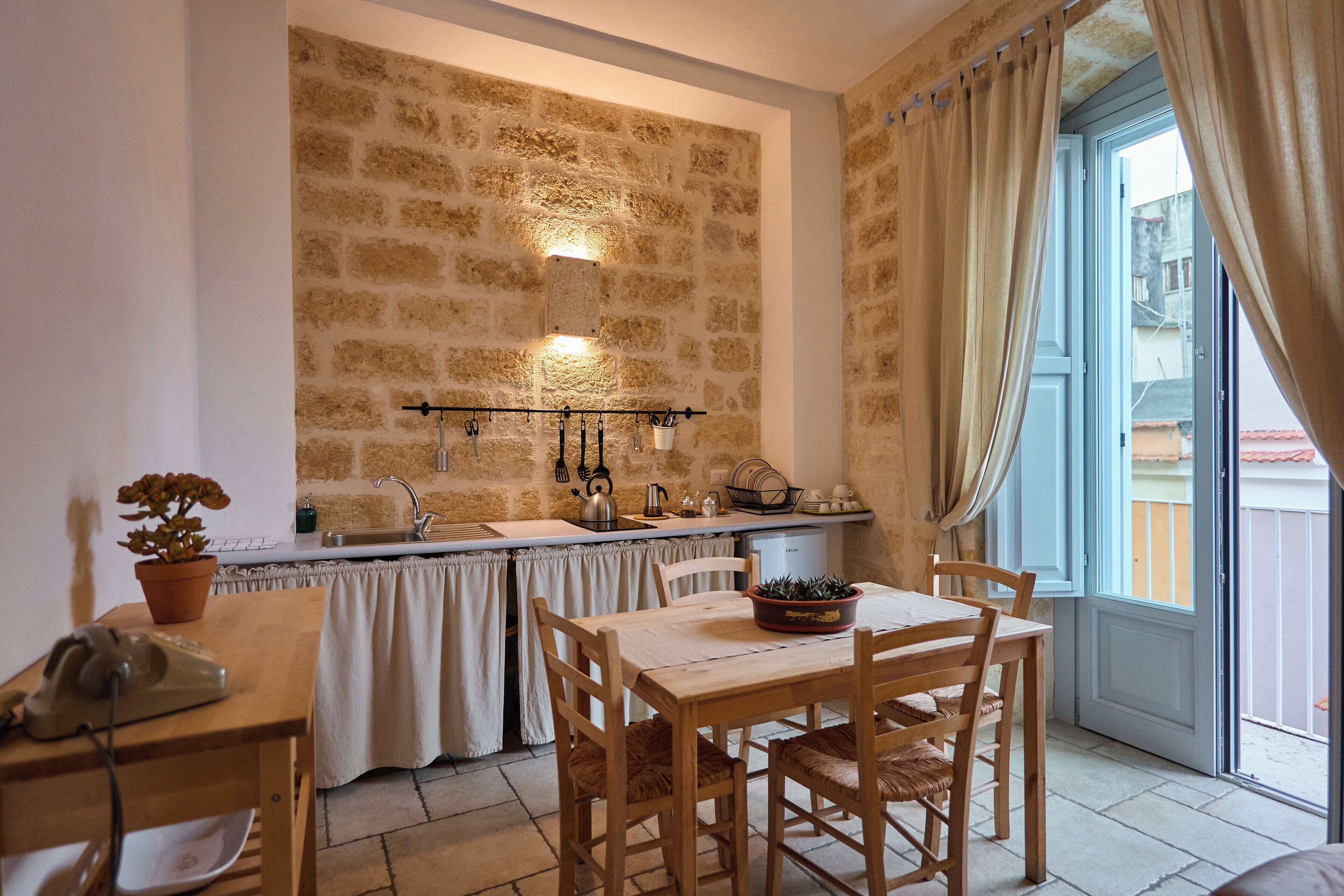 Une cuisine avec un mur en pierre, une table à manger avec des chaises, un évier et une fenêtre avec des rideaux.