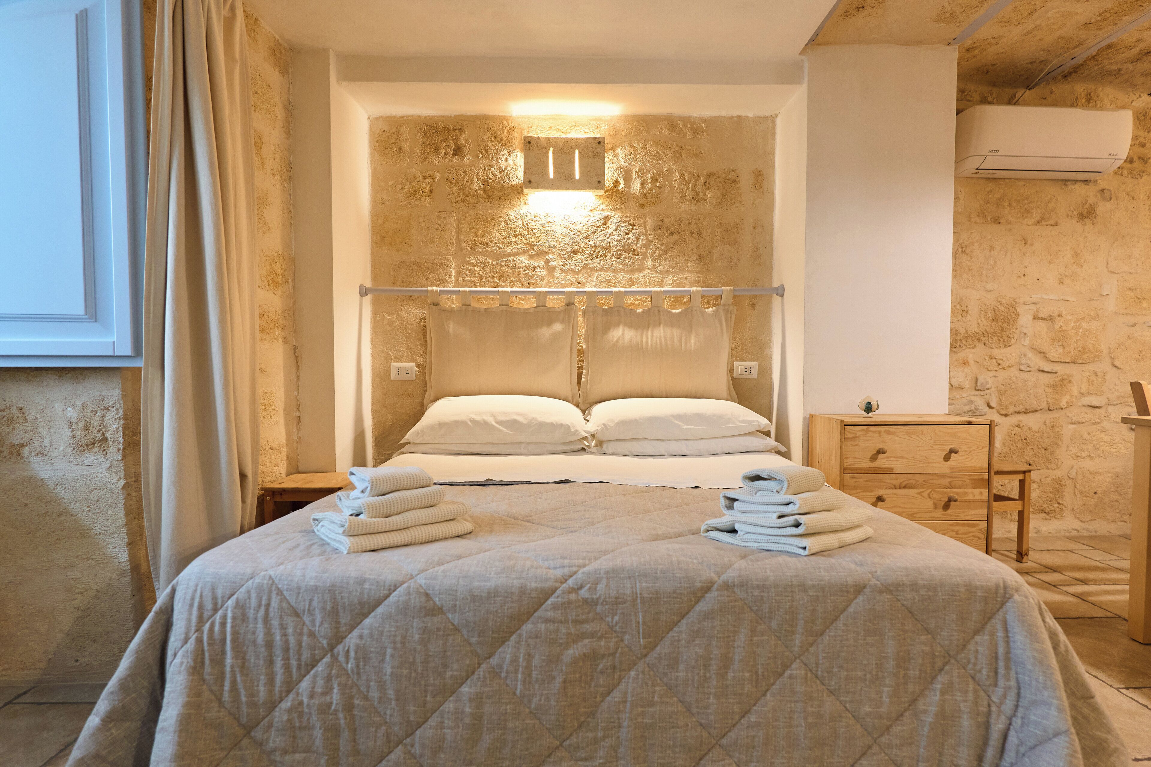 A hotel room with a large bed, a bedside table, a wooden dresser, and a stone wall.