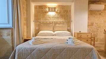 A hotel room with a large bed, a bedside table, a wooden dresser, and a stone wall.