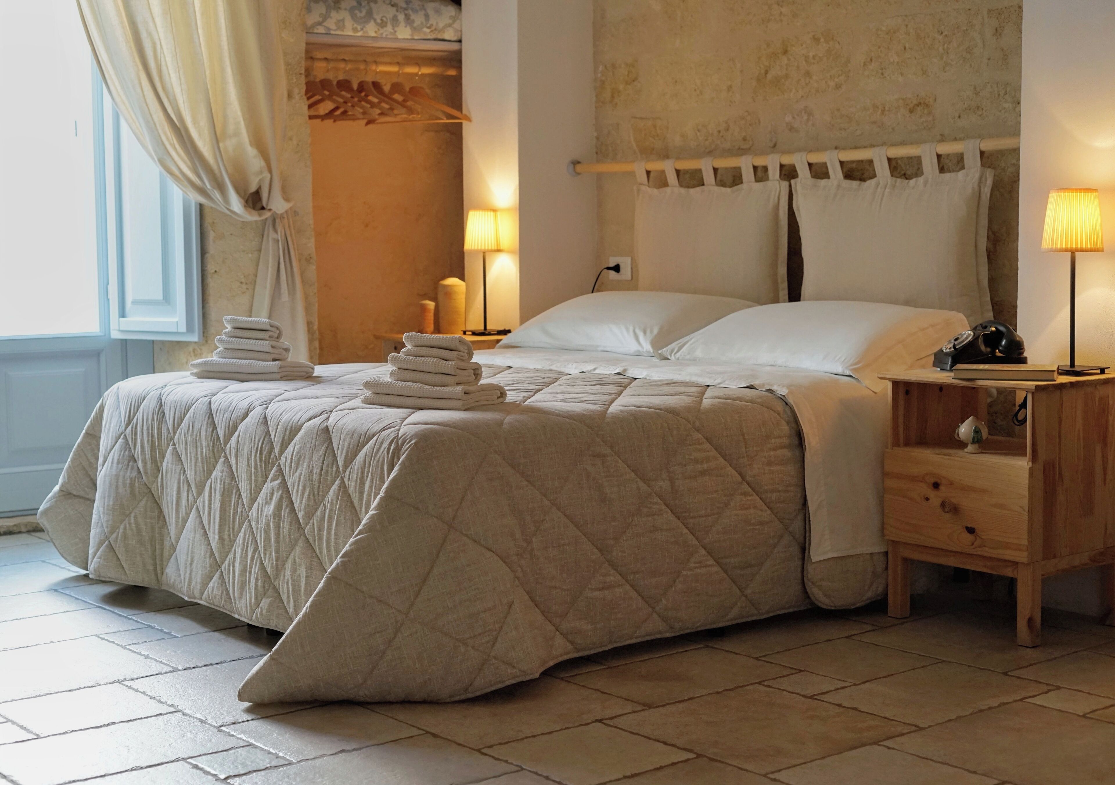 Uma cama bem arrumada com cobertura acolchoada, uma mesa de cabeceira com abajur e uma pilha de toalhas dobradas.