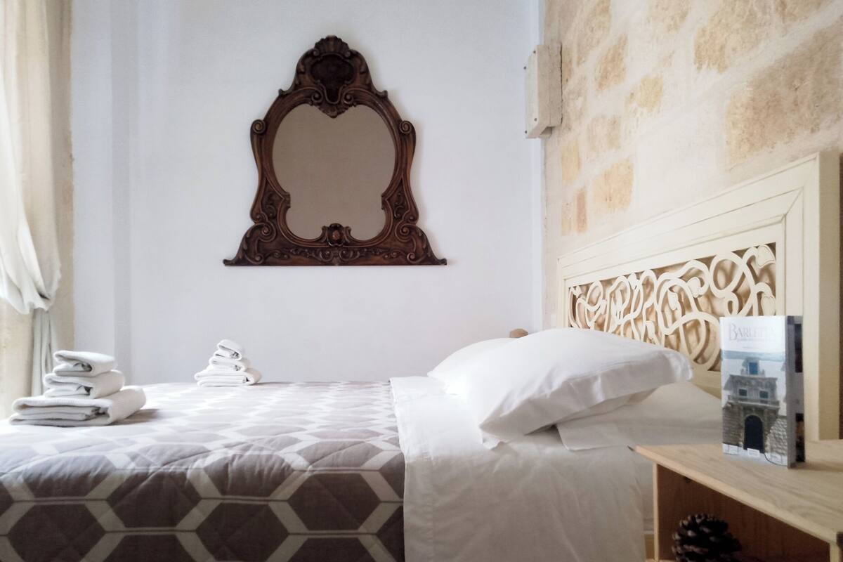 A hotel room with a bed, a headboard with intricate carvings, a wall-mounted mirror, a stack of folded towels, and a small shelf with a book and a pine cone.