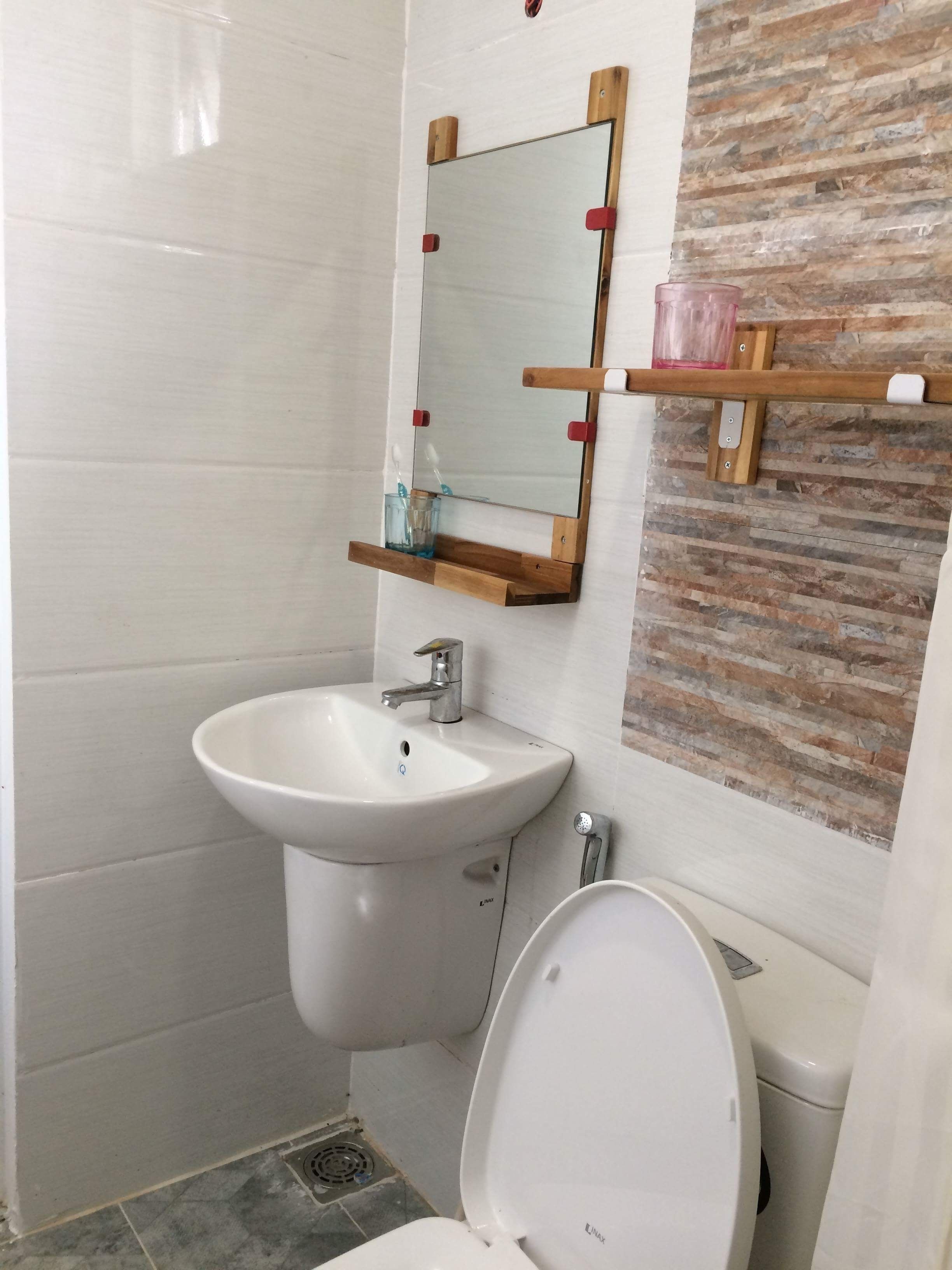 A bathroom with a white sink, a mirror, and a toilet. There is a wooden shelf with a glass container and a toothbrush holder.