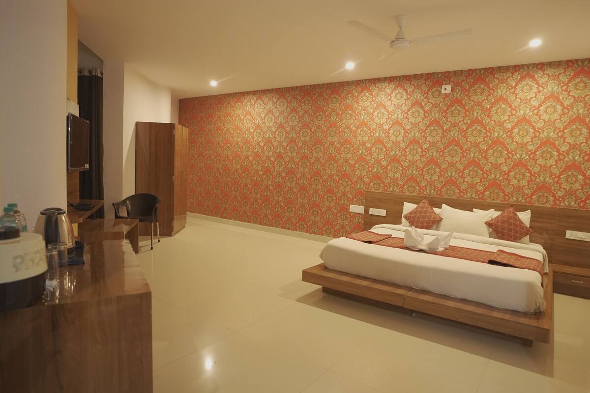 A hotel room with a large bed, a wooden headboard, a red patterned wall, a ceiling fan, and a desk with a chair.