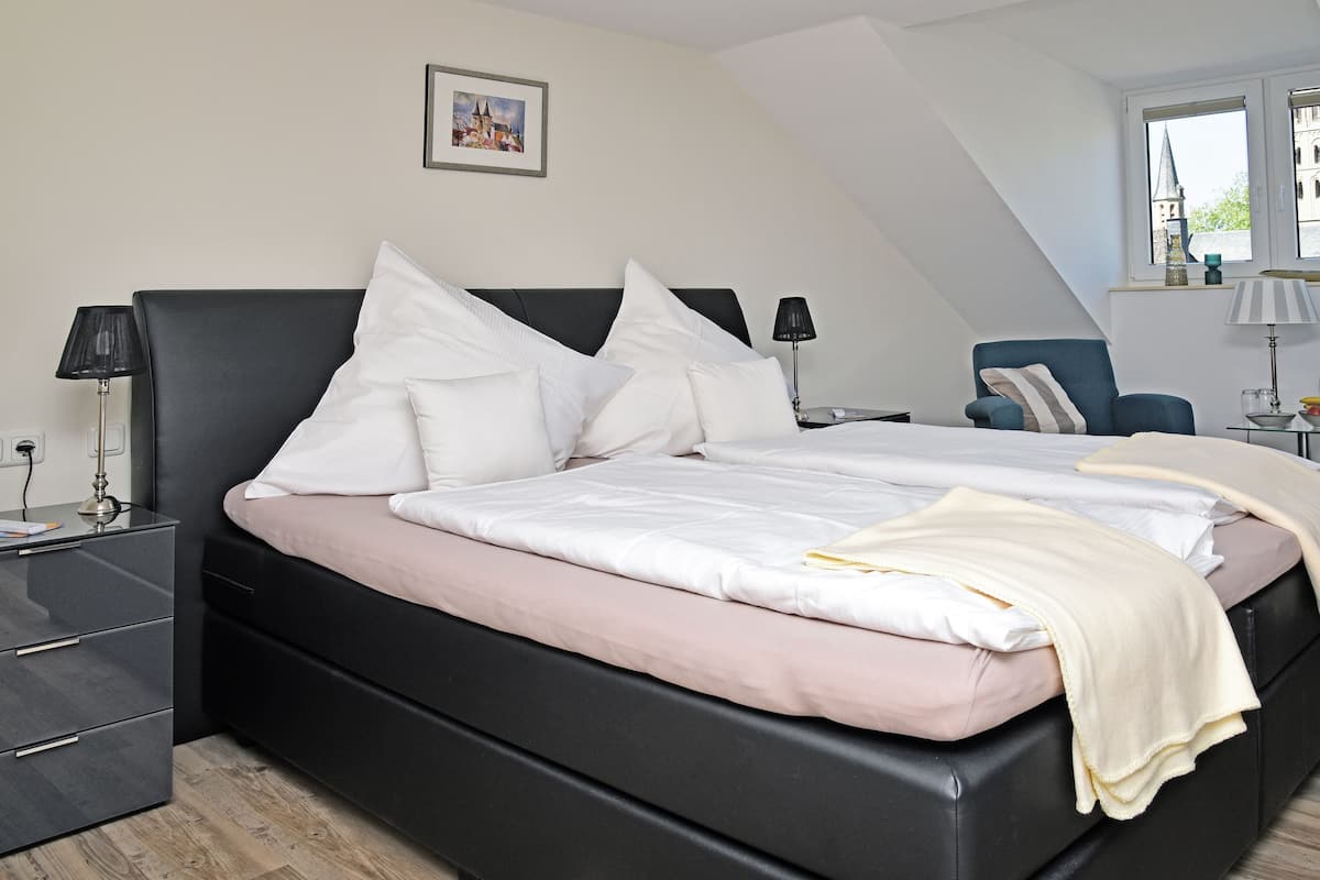 A modern bedroom with a black headboard, white bedding, a bedside table with a lamp, and a view of a building through a window.