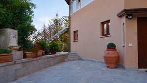 Un edificio con fachada color beige, una puerta marrón y una escalera que lleva a un nivel superior. Hay plantas en macetas y una maceta de terracota grande en el suelo.