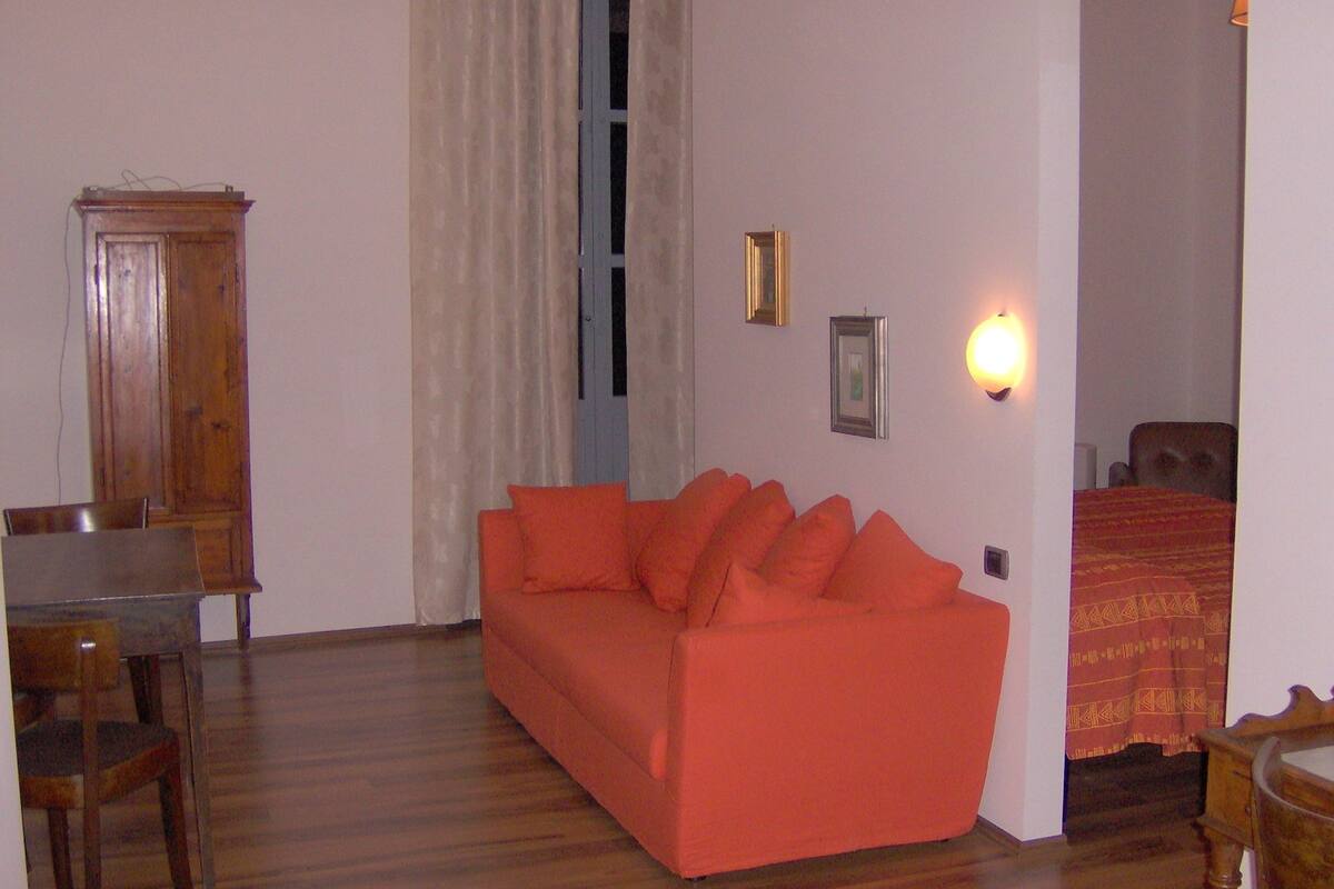 A room with a wooden floor, a red sofa, a wooden cabinet, and a bed with a patterned blanket.