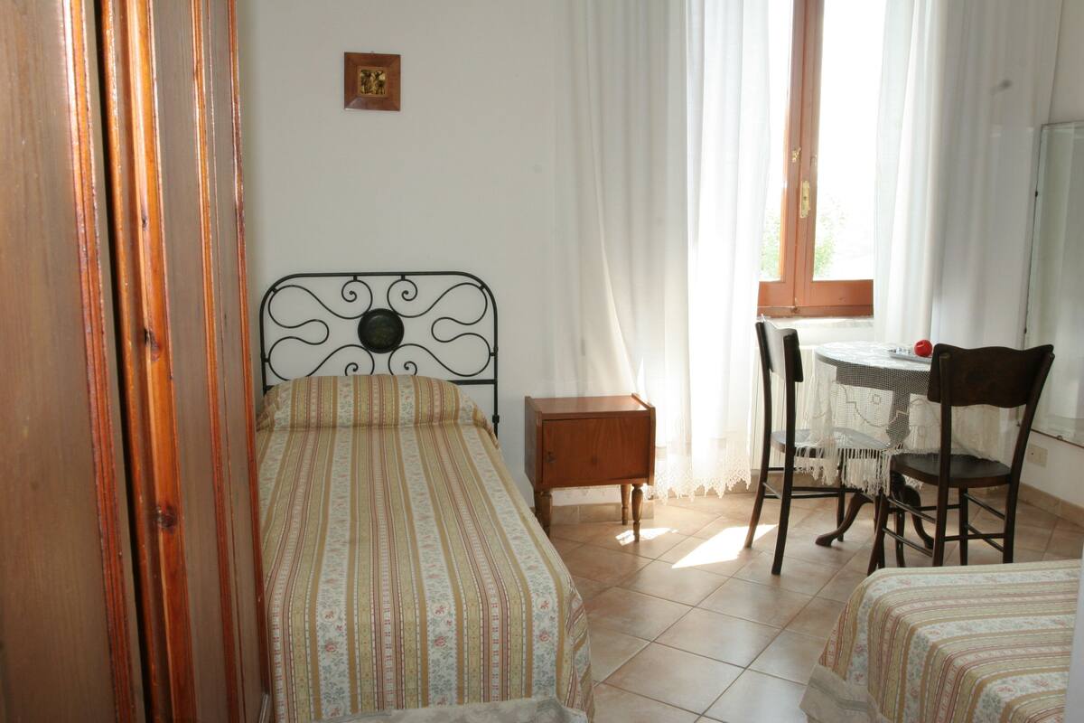 A bedroom with a bed, a small bedside table, a chair, and a desk with a chair.