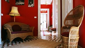 A room with a red wall, a wicker sofa with cushions, a wicker chair, a lamp, and a patterned rug. - 5 Balconi (Catania)