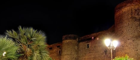 Un castillo de piedra histórico con almenas y una torre redonda, rodeado de palmeras e iluminado por faroles.