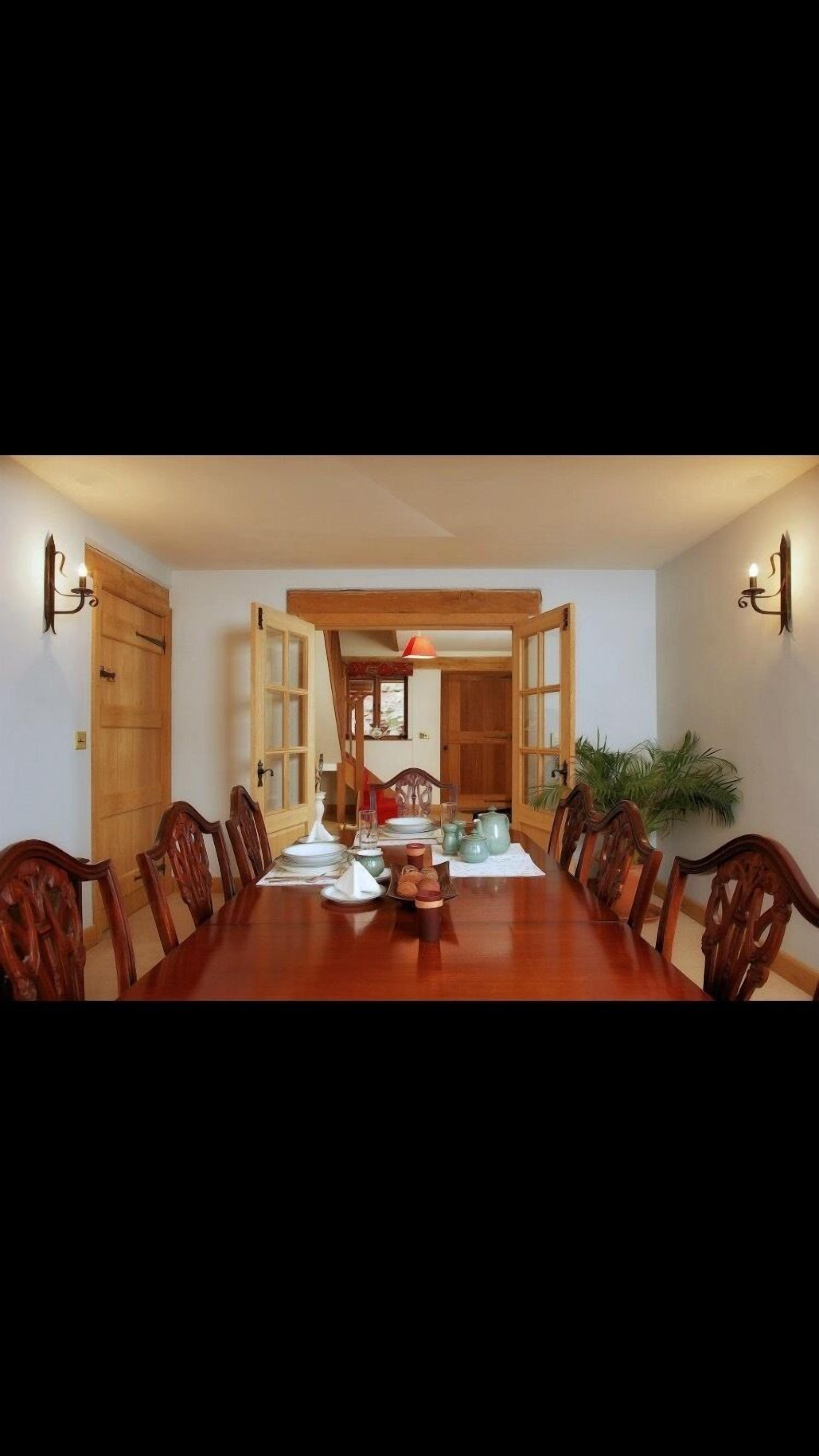 Une salle à manger avec une longue table en bois, six chaises et un fauteuil rouge. On y trouve également des appliques murales et une plante.