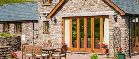 A stone building with a large wooden door and windows, a patio with a table and chairs, and potted plants.