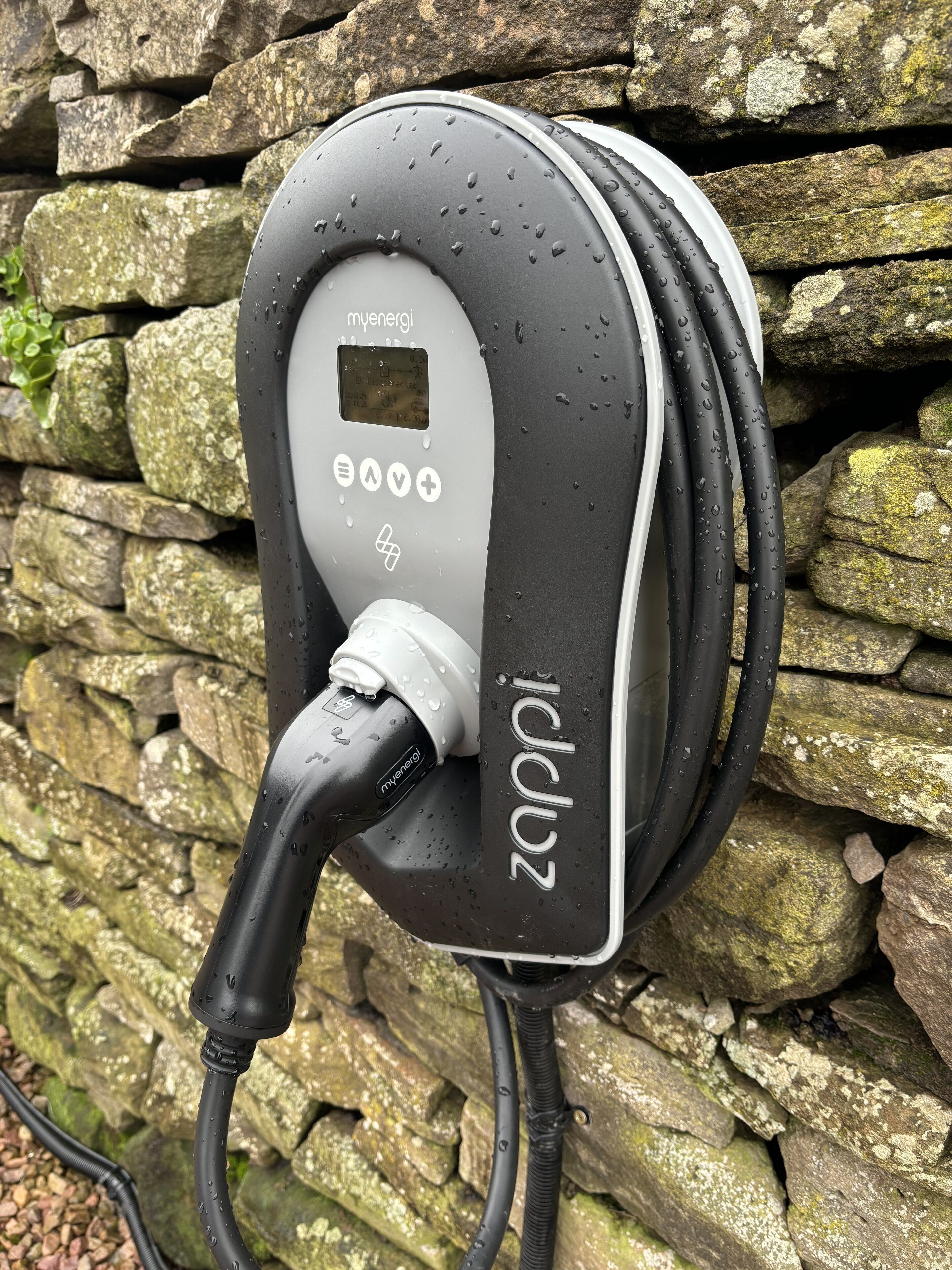 An electric vehicle charging station with a digital display, charging cables, and a stone wall background.