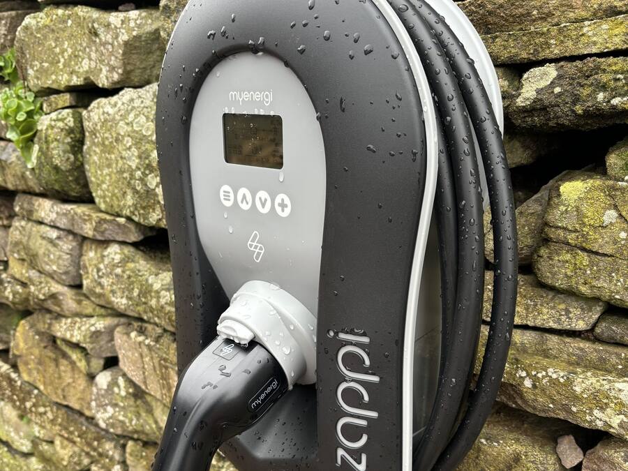 An electric vehicle charging station with a digital display, charging cables, and a stone wall background.