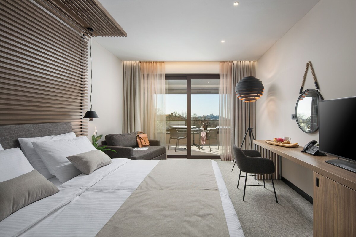 A modern hotel room with a large bed, a desk with a chair, a TV, and a balcony with a table and chairs.