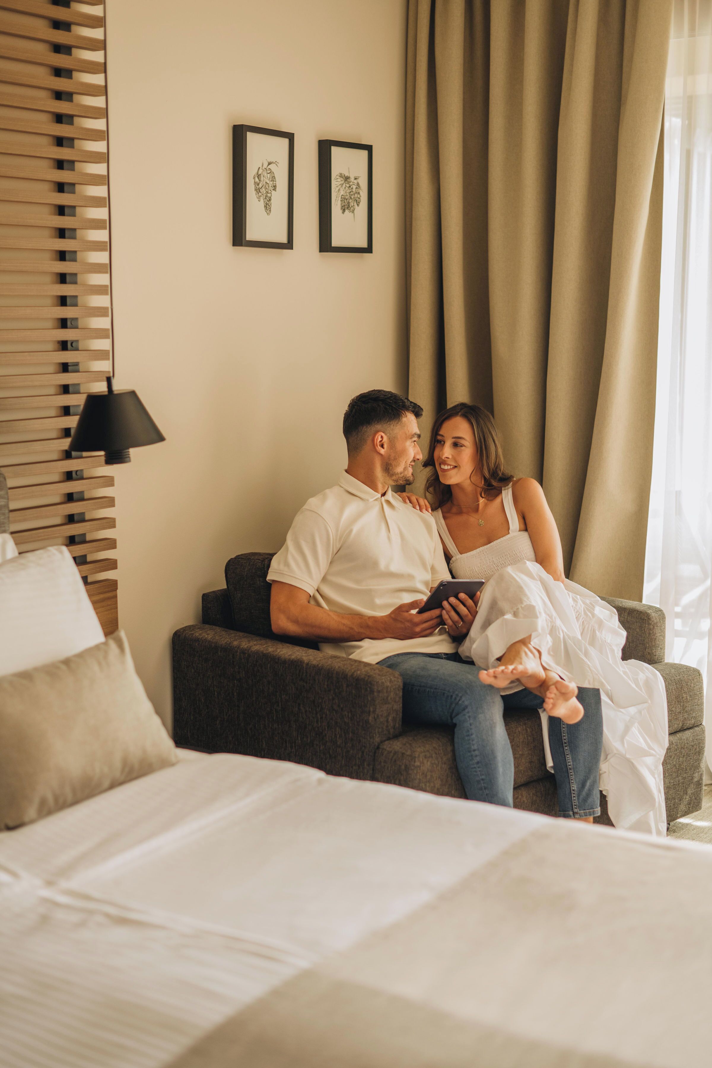 Una pareja sentada en un sofá en una habitación de hotel, con una cama, una lámpara y cortinas.