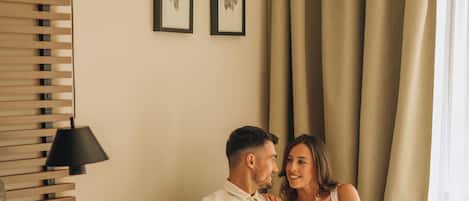 Una pareja sentada en un sofá en una habitación de hotel, con una cama, una lámpara y cortinas.