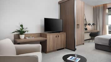 A modern hotel room with a sofa, a coffee table with a magazine, a TV on a wooden cabinet, a desk with a laptop, and a bed.