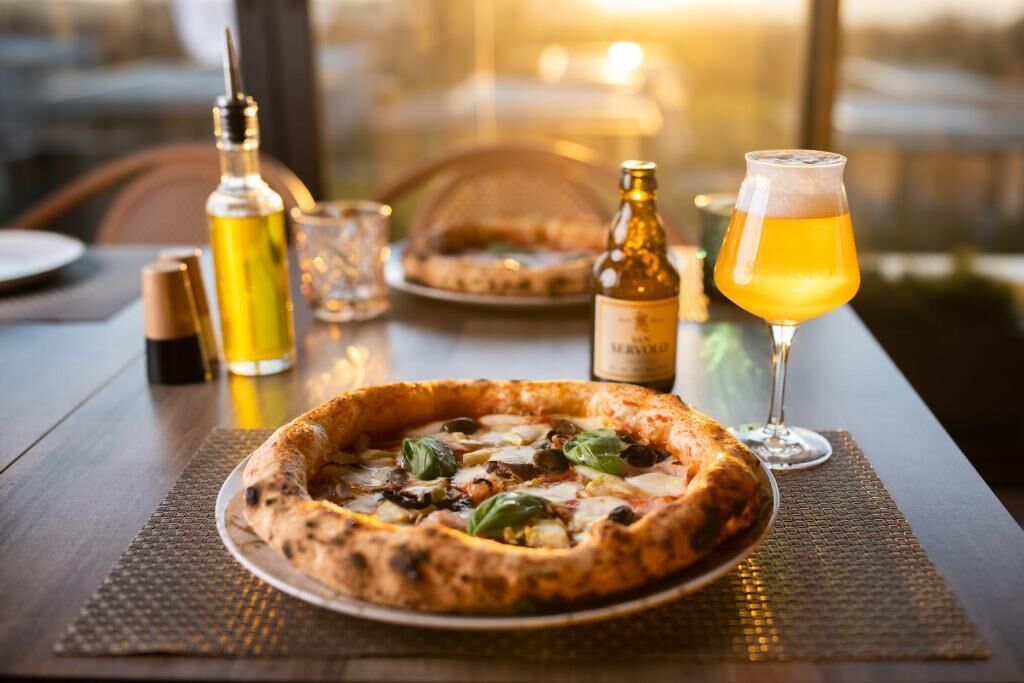 A pizza on a plate with a glass of beer and a bottle of olive oil on a table.