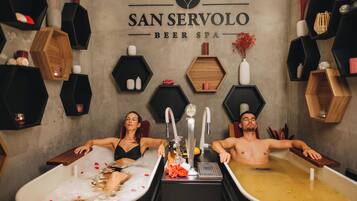Two people relaxing in separate tubs at a spa, with a sign reading "SAN SERVOLO BEER SPA" on the wall.