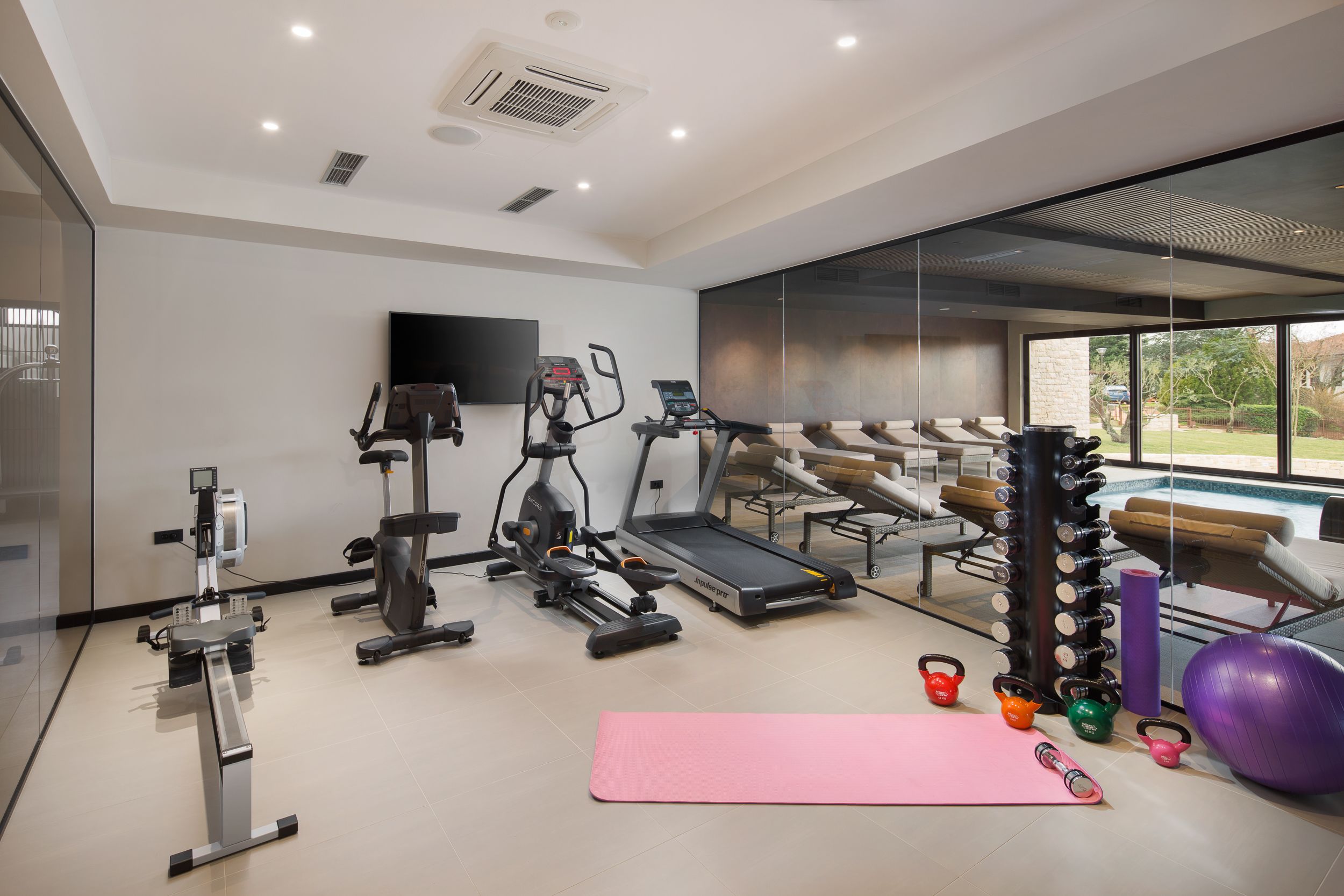 A modern gym with cardio machines, a yoga mat, kettlebells, and a large window offering a view of outdoor seating.