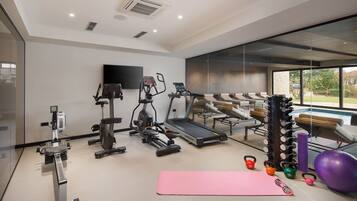 A modern gym with cardio machines, a yoga mat, kettlebells, and a large window offering a view of outdoor seating.