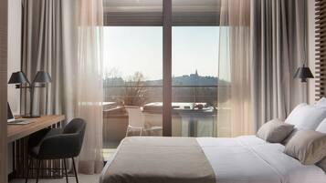 Habitación de hotel con cama, escritorio, silla y vistas a un paisaje urbano a través de amplias ventanas.