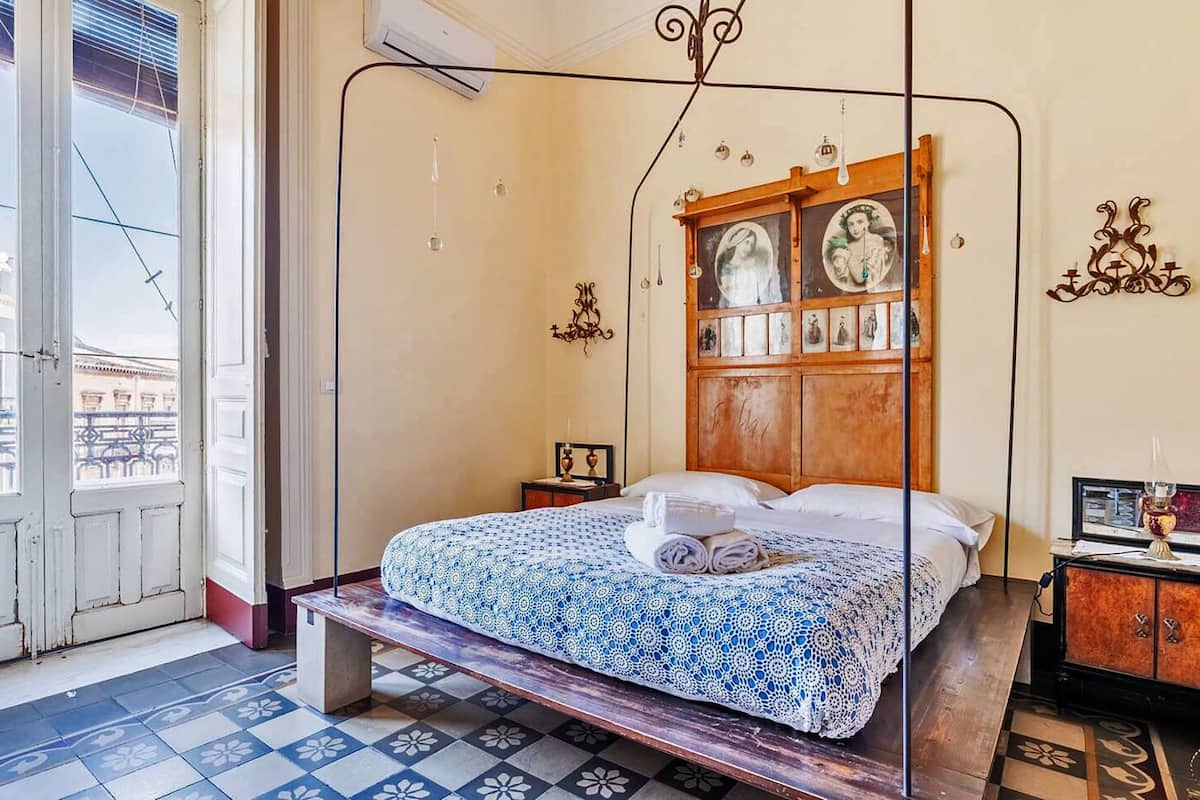 A room with a four-poster bed, a wooden headboard with artwork, a tiled floor, and a view through an open door.