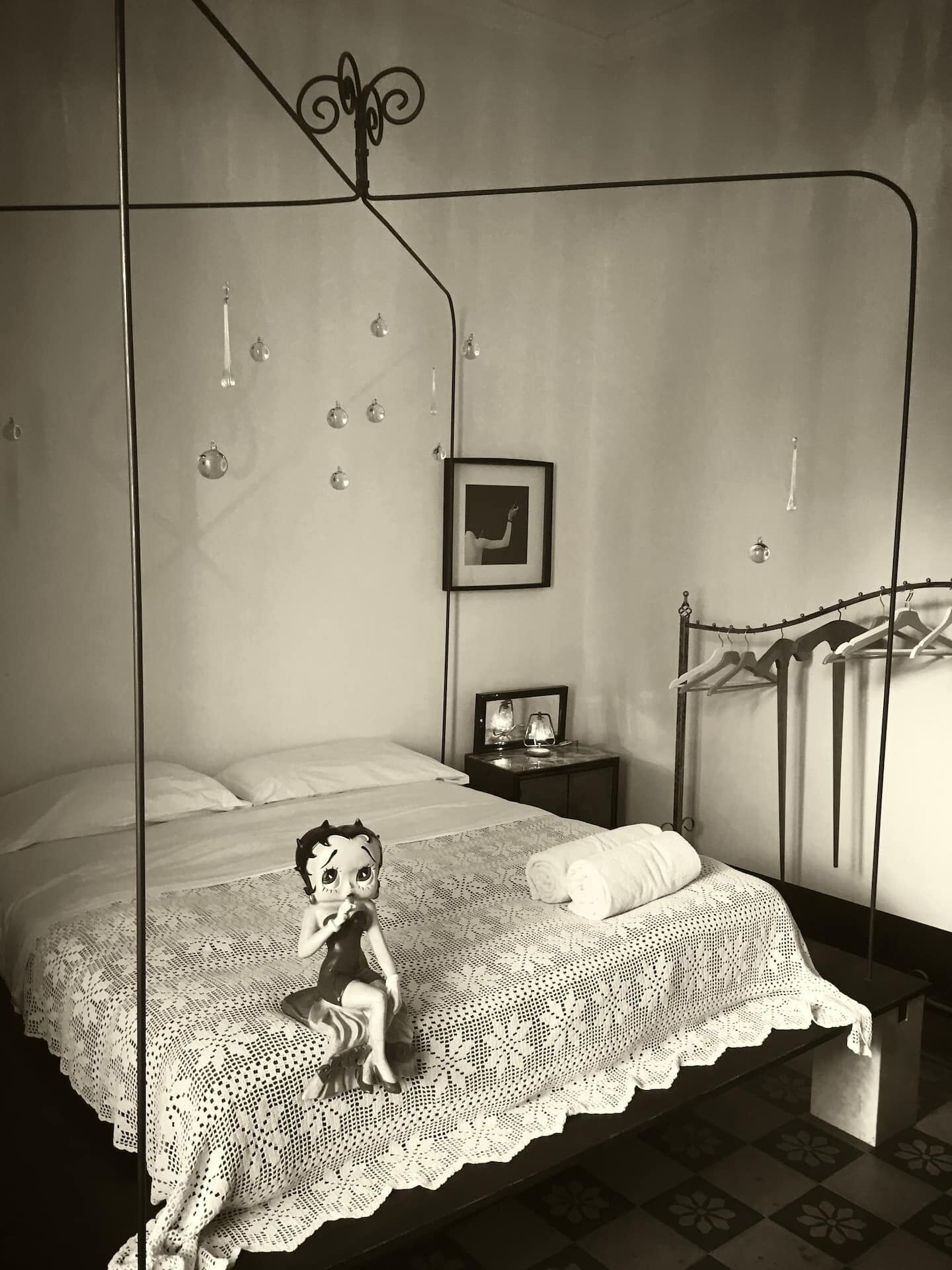 A bedroom with a four-poster bed, a small bedside table with a lamp, a framed picture on the wall, and a hanging light fixture.