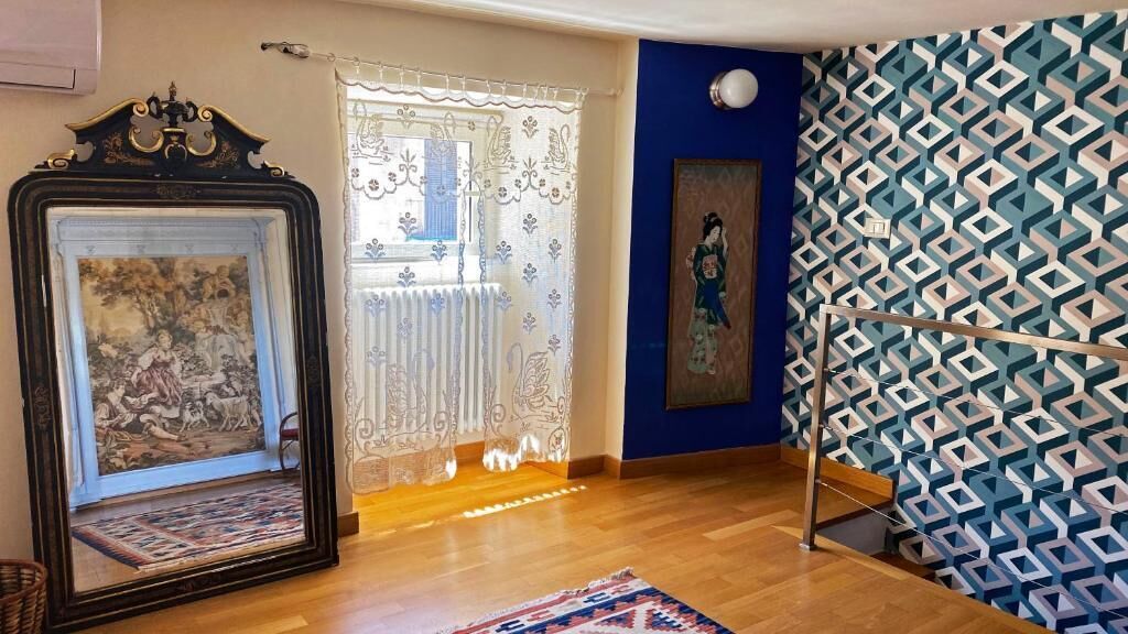 Een kamer met een houten vloer, een grote spiegel met decoratieve lijst, een gordijn met een patroon en een blauwe muur met een geometrisch ontwerp.