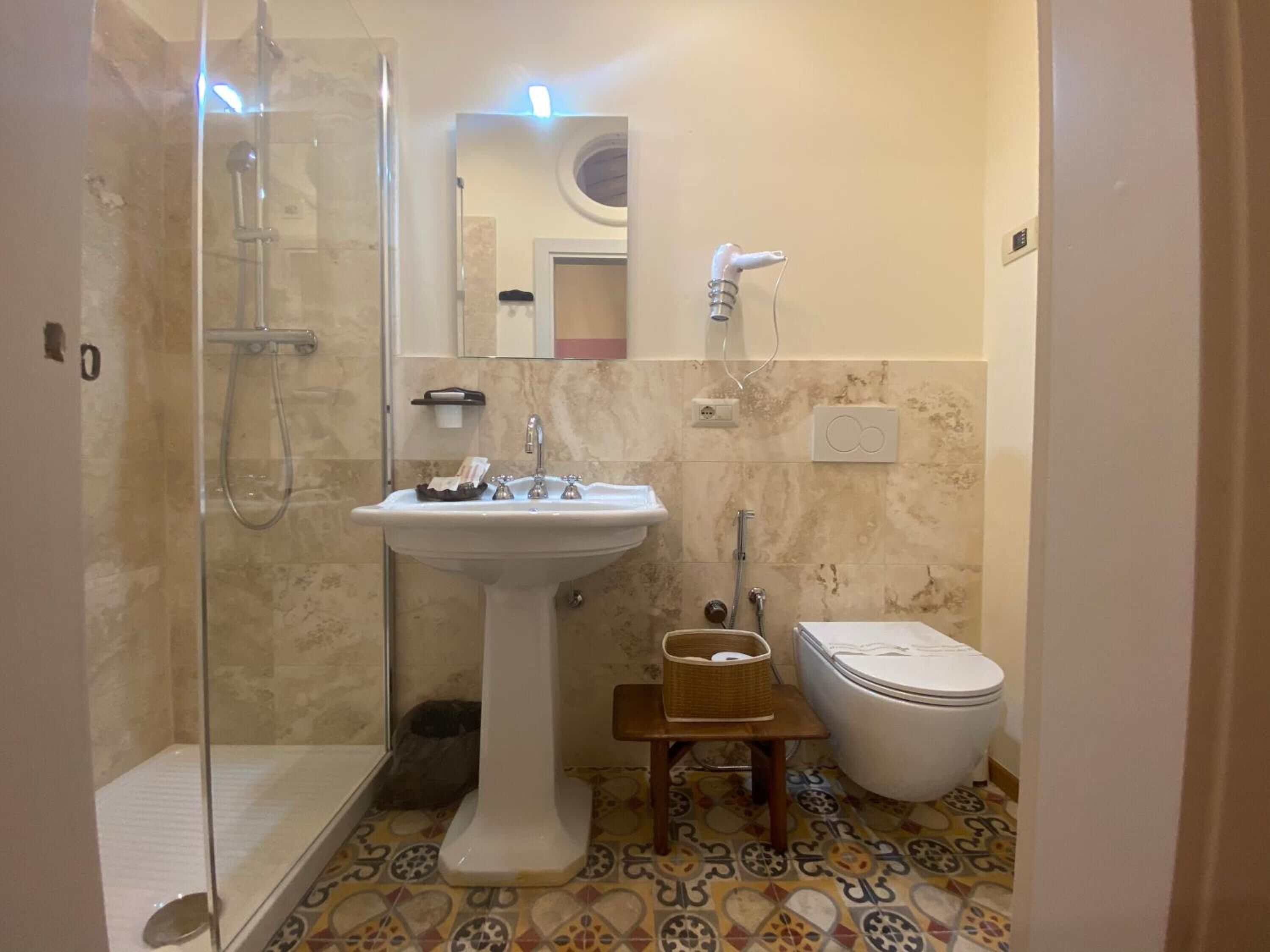 A bathroom with a glass shower enclosure, a pedestal sink, a toilet, and a small wooden stool.