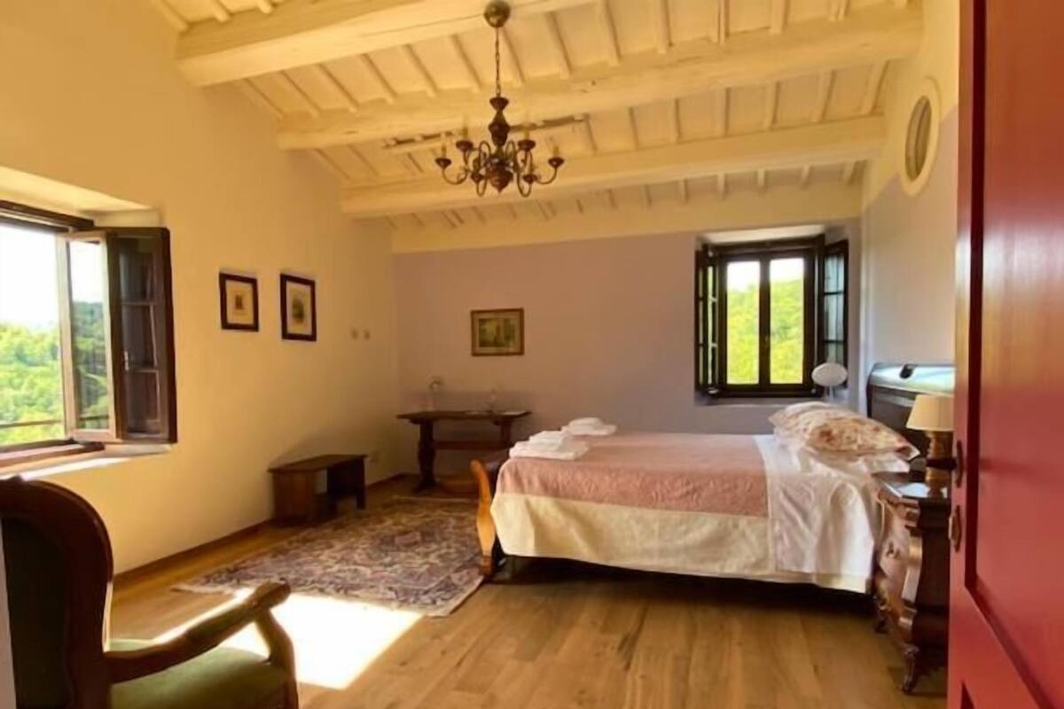 A bedroom with a wooden floor, a bed with white bedding, a desk, a chair, and a chandelier.