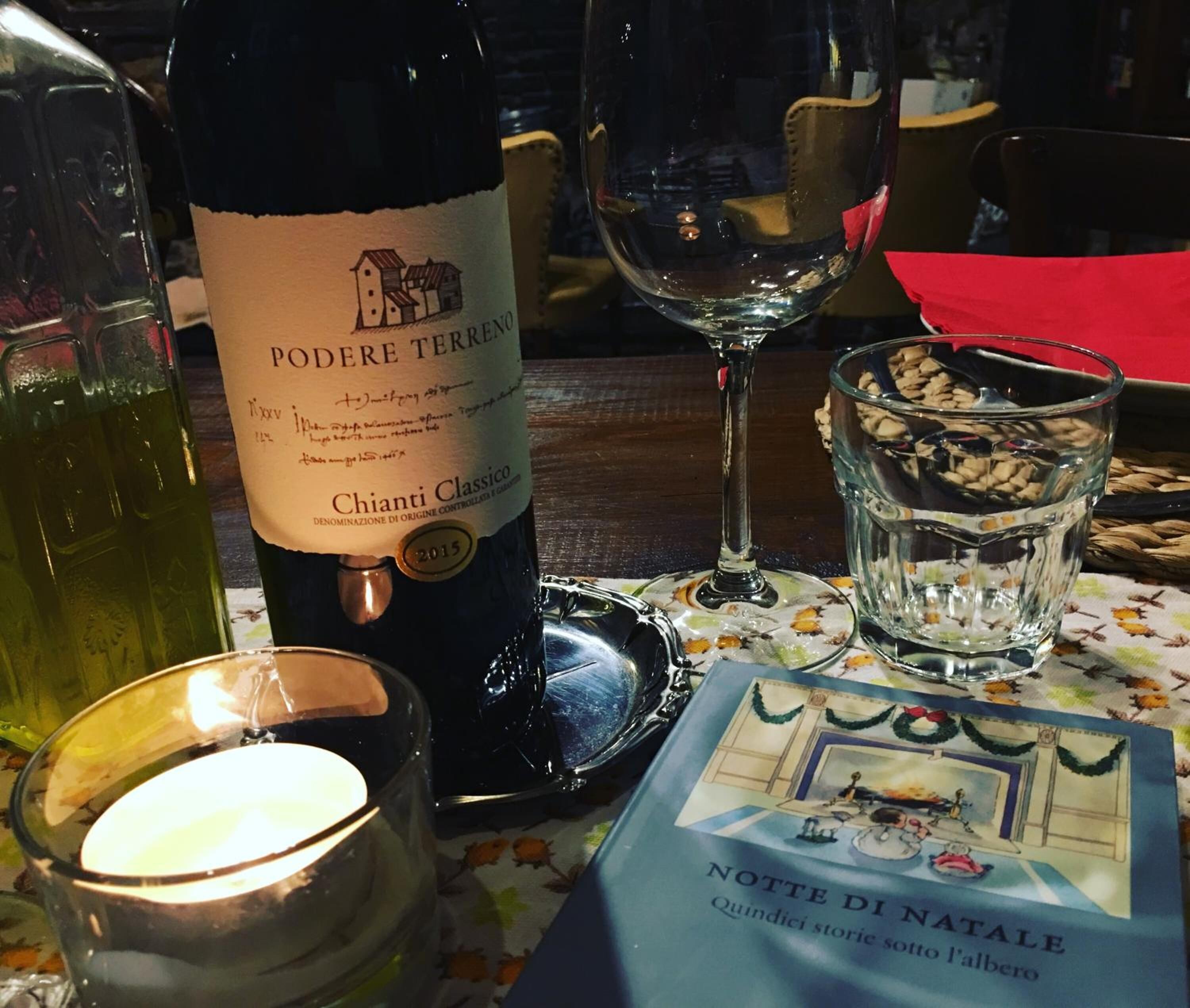 A bottle of Chianti Classico wine, a glass of wine, a candle, and a book titled "Notte di Natale" on a table.