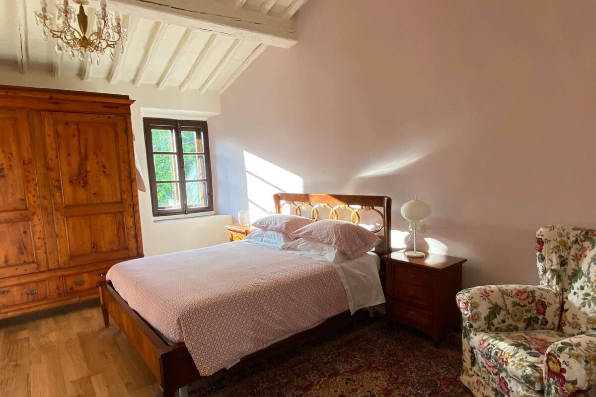A bedroom with a wooden bed, a nightstand, a chair, a chandelier, and a window with a view of greenery.