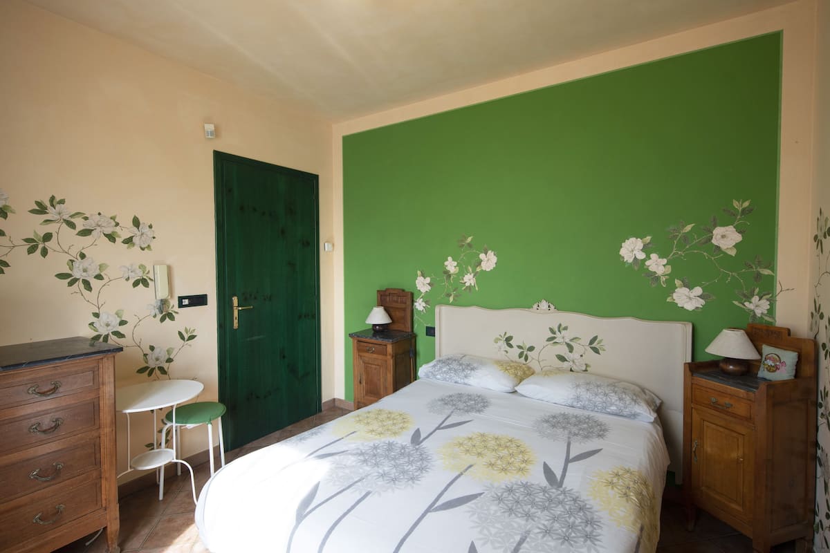 Une chambre avec un mur d’accent vert, un lit avec une literie à motifs floraux, une table de chevet, une commode et un petit tabouret.