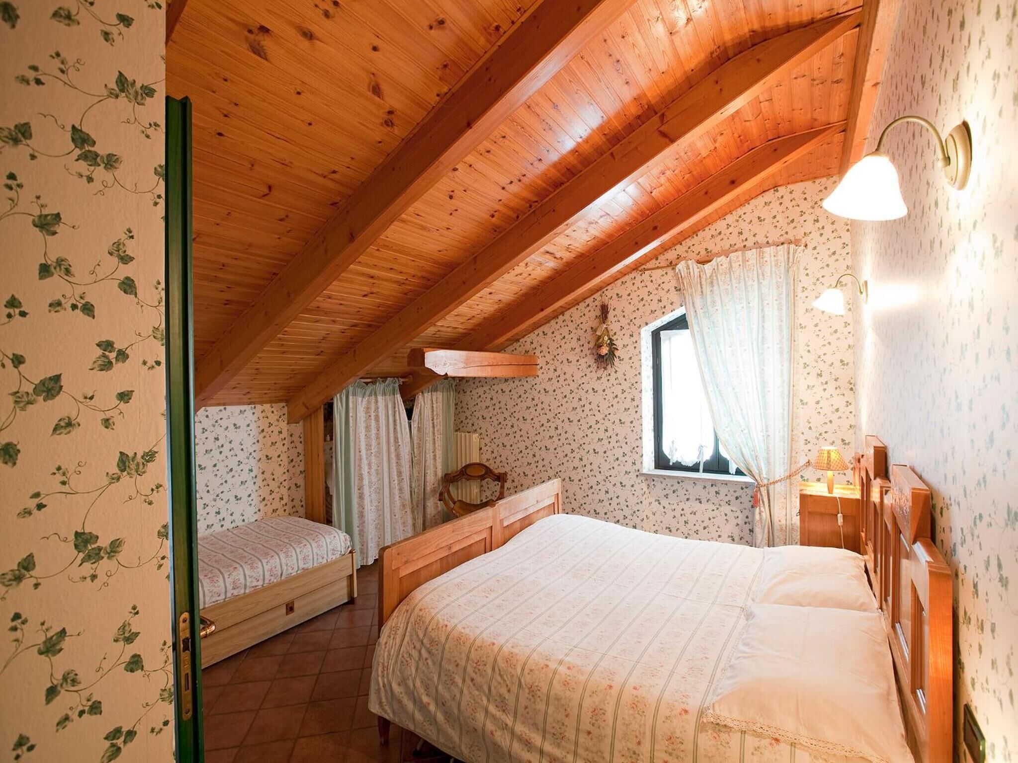 A cozy attic bedroom with wooden beams, two beds, floral wallpaper, and a window with curtains.