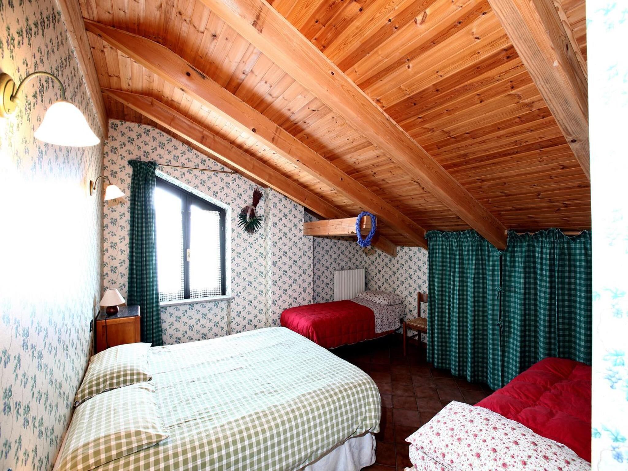 A cozy attic bedroom with two beds, wooden ceiling beams, floral wallpaper, and a window with curtains.
