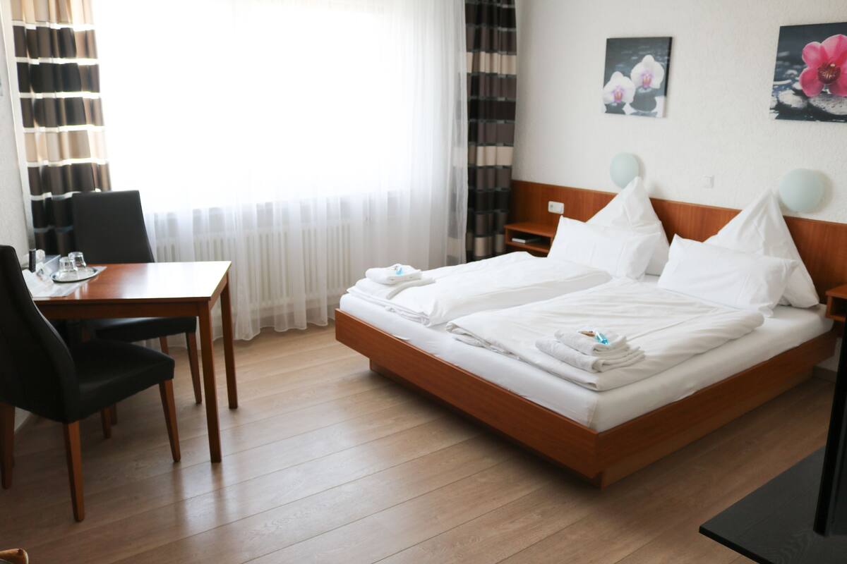 Une chambre d’hôtel avec un lit en bois, un bureau avec une chaise et une fenêtre ornée de rideaux légers.