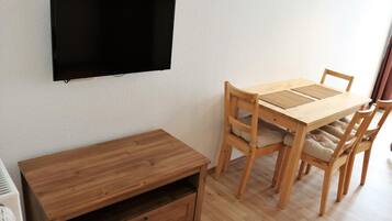 A room with a wooden dining table, four chairs, a TV mounted on the wall, and a small wooden cabinet.