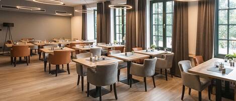 Un vaste espace dédié à la salle à manger, avec de nombreuses tables et chaises, de grandes fenêtres dotées de rideaux et des luminaires circulaires modernes au plafond.