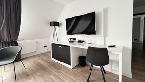 A modern hotel room with a white desk, two chairs, a flat-screen TV, a small cabinet, and a floor lamp. -  DORMERO Hotel Xanten (Xanten)