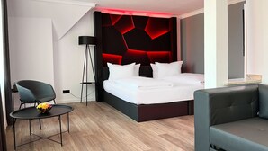 A modern hotel room with a bed, a chair, a small table, and a floor lamp. -  DORMERO Hotel Xanten (Xanten)