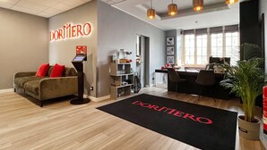 A hotel lobby with a reception desk, a sofa with red cushions, a coffee machine, and a large window. -  DORMERO Hotel Xanten (Xanten)