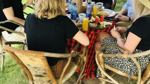 Un grupo de personas sentadas en una mesa al aire libre, con diferentes comidas y bebidas, bajo una estructura de techo de paja.
