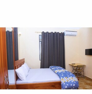 A bedroom with a single bed, a small table, a chair, a wall-mounted air conditioner, and a curtain. - Chibuba Airport Accomodation (Dar es Salaam)