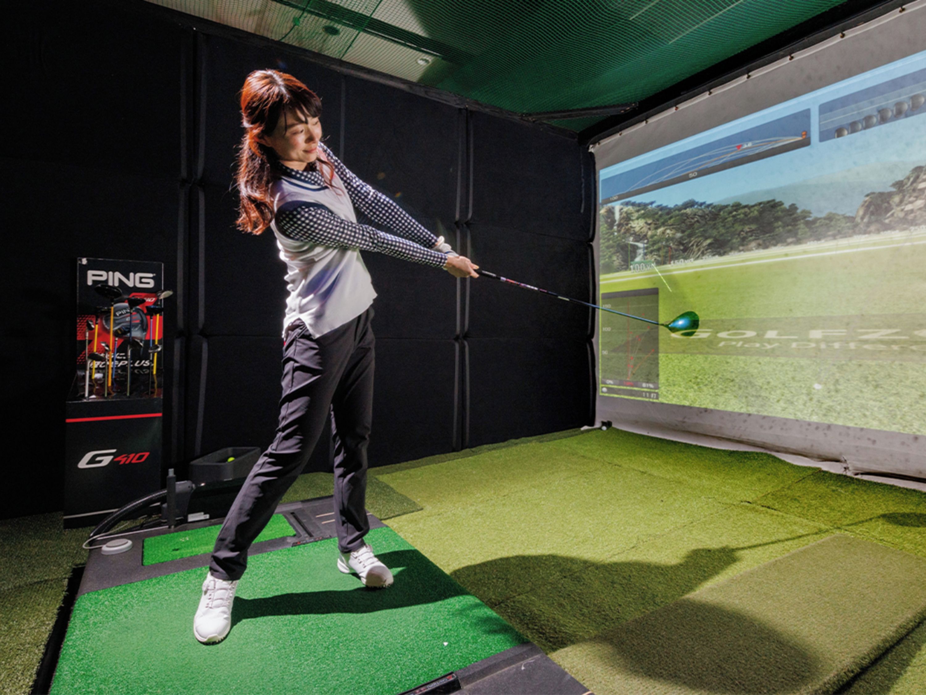 Una persona practicando golf en un campo de práctica con una pantalla digital que muestra una vista de un campo de golf.