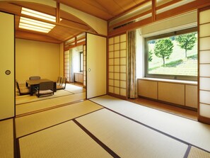 Una habitación japonesa tradicional con suelo de tatami, una mesa baja con cojines, puertas correderas y vistas a un camino bordeado de árboles.