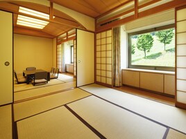 Una habitación japonesa tradicional con suelo de tatami, una mesa baja con cojines, puertas correderas y vistas a un camino bordeado de árboles.