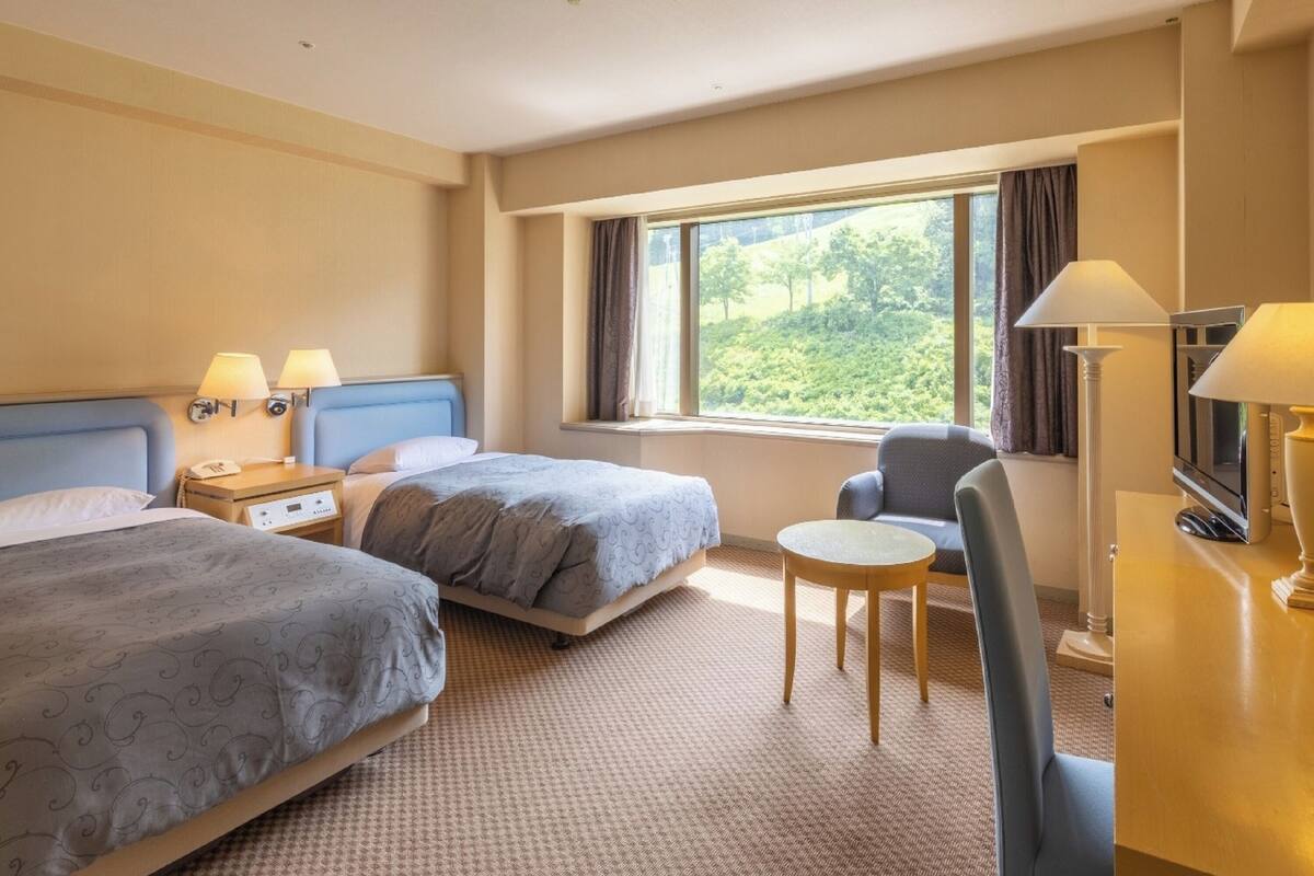 Une chambre d’hôtel avec deux lits, un bureau, une chaise, une télévision et une fenêtre donnant sur un paysage verdoyant.